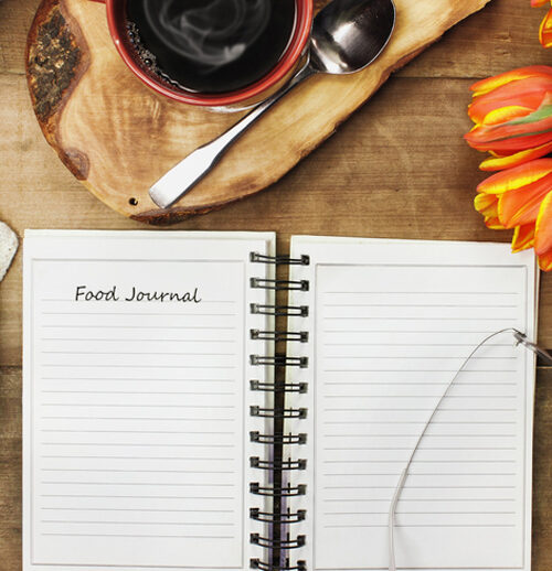 Overhead shot of an open food journal book with cell phone, coffee and flowers over a wood table top ready to plan diet. Flat lay top view style.; Shutterstock ID 576134338; Purchase Order: GSK03042019